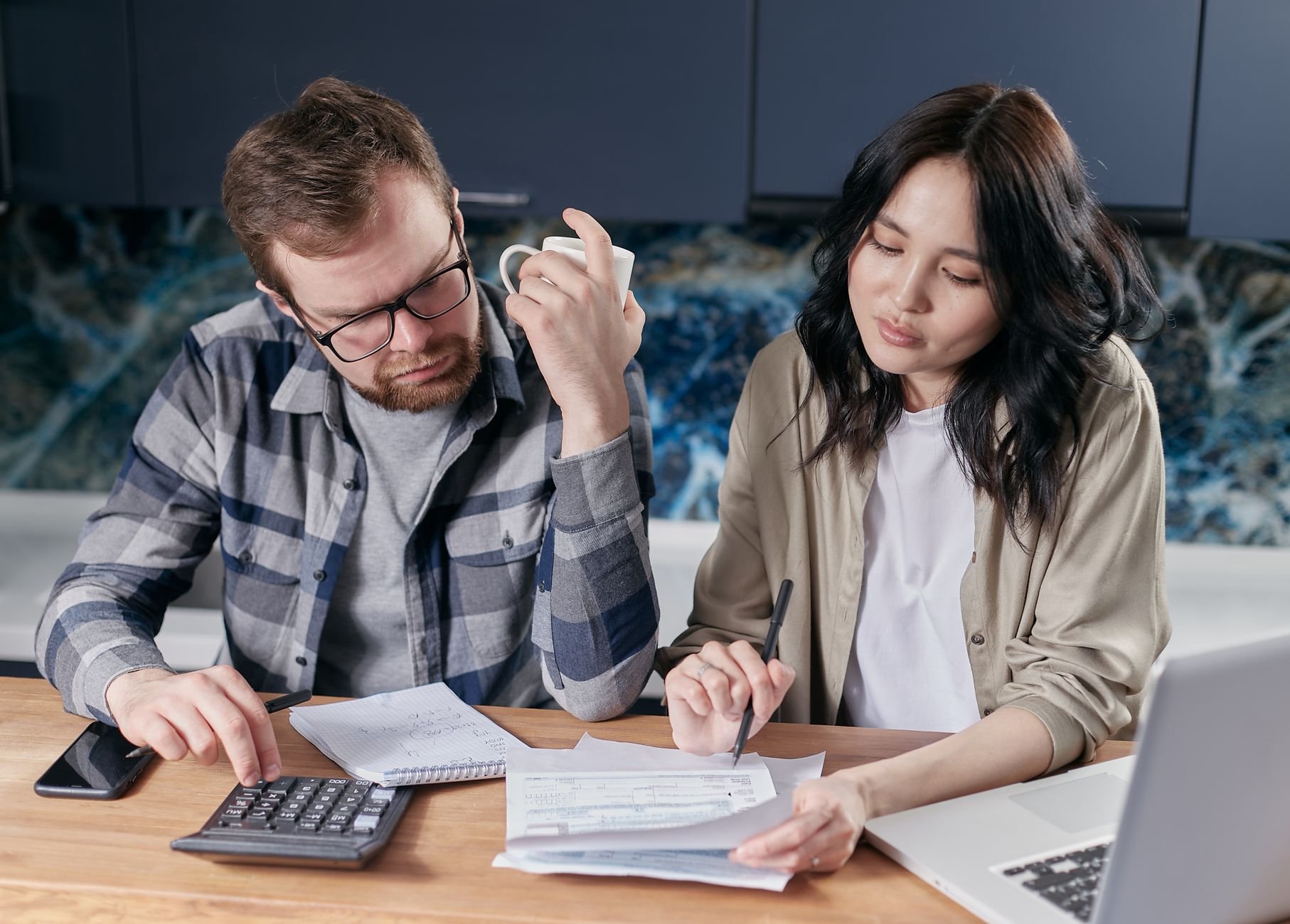 A couple reviewing household bills and budget using a calculator and laptop at their kitchen table